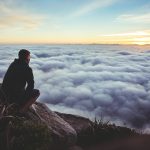 man sitting on gray rock while staring at white clouds