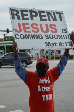 Man in red shirt holds a sign with religious message at a busy intersection.