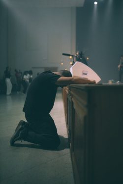 A man kneels in silent prayer during a concert in a dimly lit room.