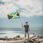 man in blue t-shirt holding flag standing on brown rock near body of water during
